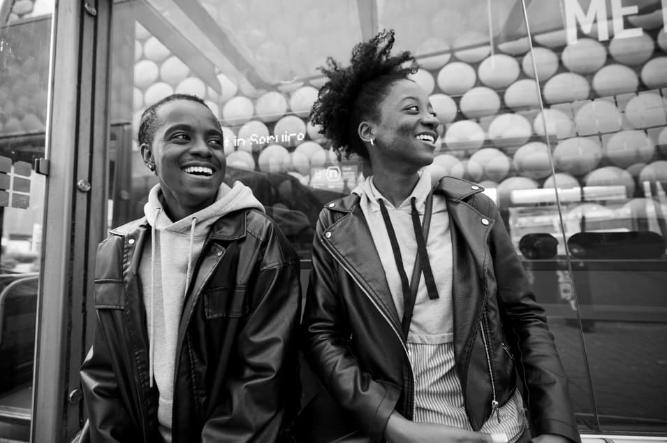 Two individuals wearing leather jackets over hoodies, standing side by side in front of a bus stop