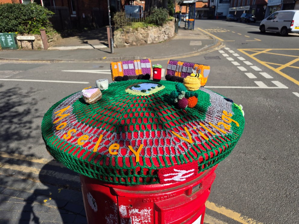 A knitted topper on top of a red post box. There is a knitted West Midlands Railway train in the centre, as well as others details like knitted fruit and vegetables. The words 'Moseley Village' are knitted around the edge.