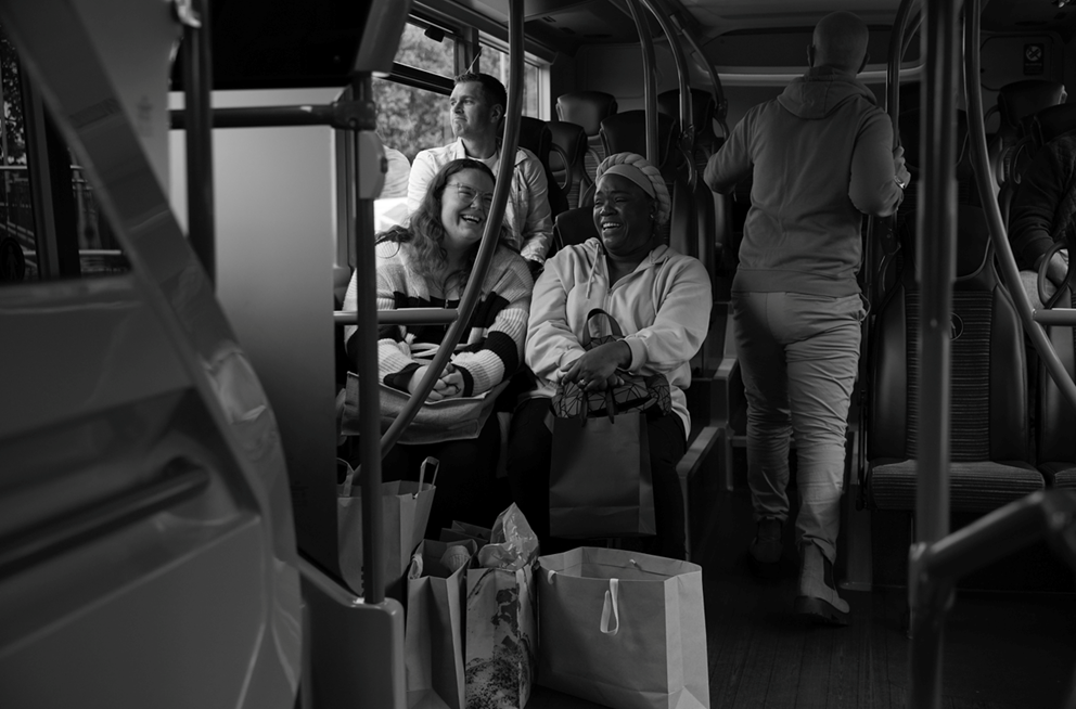 An image of two people happily riding the bus. They have lots of shopping bags stored next to their seats.
