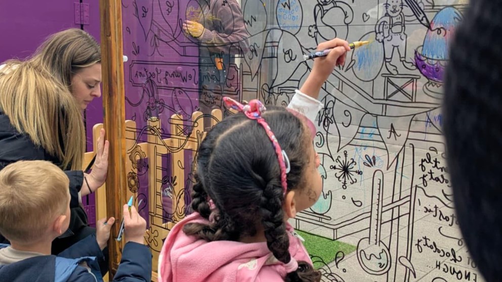Children and an adult drawing cartoon characters and balloons on a glass wall with colorful markers, with a wooden fence and greenery in the background