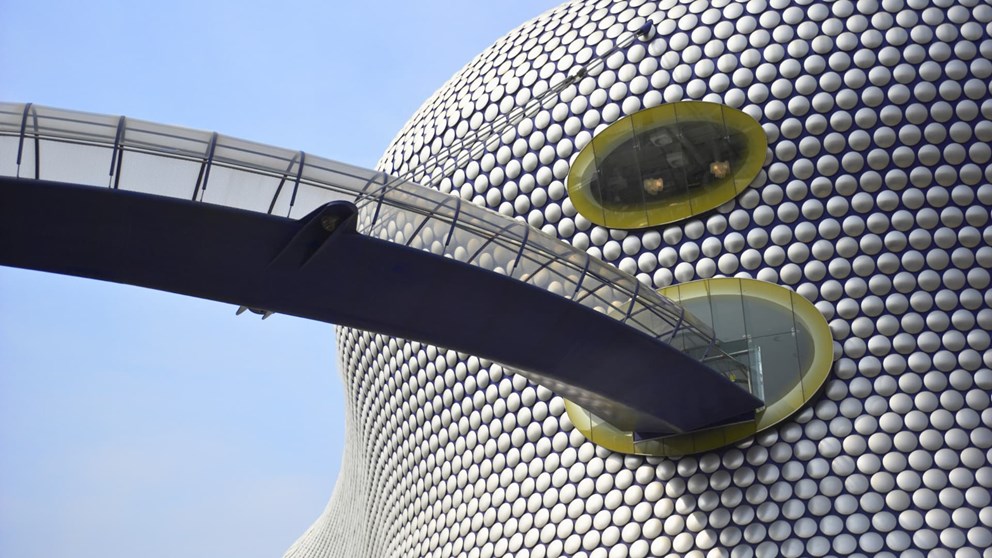 Modern building with circular metallic discs, oval yellow-bordered windows, and a curved glass pedestrian bridge against a clear blue sky